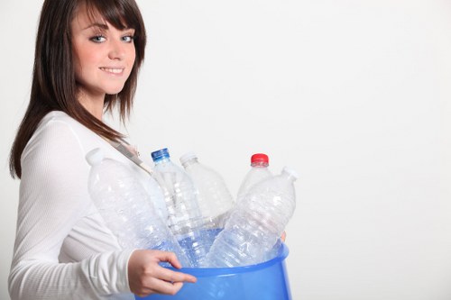 A young woman with shoulder-length brown hair, dressed in a white long-sleeved top, is smiling softly while holding a blue plastic bin filled with multiple discarded clear plastic bottles, some with red and blue caps. She stands against a plain white background, and the bottles in the bin are of various sizes, appearing crumpled and used. The scene suggests the collection or disposal of recyclable plastic waste, relevant to rubbish removal services in the Hillingdon area, as promoted by companies like Office Clearance Hillingdon. The focus is clearly on the bottles and the bin, with no other objects in view. The lighting is bright, evenly illuminating the subject and the contents of the bin, emphasizing the clarity and texture of the plastic bottles. This image reflects waste sorting and recycling themes, aligning with rubbish collection and environmental management activities common in London postcode districts near Hillingdon.