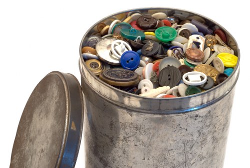 A cylindrical metal bin filled with a variety of plastic buttons of different sizes, colors, and finishes, including black, white, blue, red, yellow, and brown, some with four holes and others with two or decorative shapes. The bin is situated outdoors on a paved surface, with its round metal lid lying to the left, partly visible and resting against the bin. The metal surface of the bin appears slightly weathered with minor scratches and marks. The background is plain and white, emphasizing the bin and its contents, aligning with rubbish removal services that handle small discarded items like buttons, often found during office or domestic clearance in Hillingdon or nearby areas.