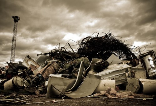 A large pile of discarded metal and household appliances, including washing machines, refrigerators, and assorted scrap metal, is situated outdoors on a dirt surface. The objects are damaged, with visible dents, rusted surfaces, and broken parts. The pile is irregular and appears to be hastily assembled, with some items leaning or stacked haphazardly on top of others. Behind the debris, there is a tall metal tower or mast extending upward, against a cloudy, overcast sky that casts a muted, diffuse light across the scene. The surrounding environment suggests an informal or industrial waste site, typical of rubbish clearance activities, potentially serving areas like Hillingdon or nearby locations. This image, associated with a rubbish removal service such as Office Clearance Hillingdon, captures the type of industrial scrap and household waste that professional disposal services manage for commercial and domestic clients in the local area.