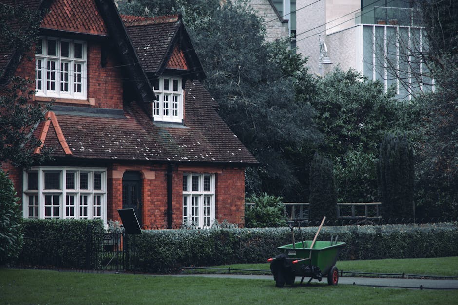 A small, red-brick house with a steep, tiled roof and white-framed windows is situated in a garden area. The house features two prominent dormer windows on the upper level and a central front door pai
