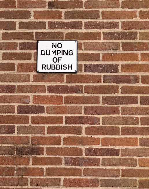A collection of discarded household items and debris piled along an outdoor brick-paved area near a wall. The scene includes a wooden pallet leaning against a small, damaged cabinet with a partially b