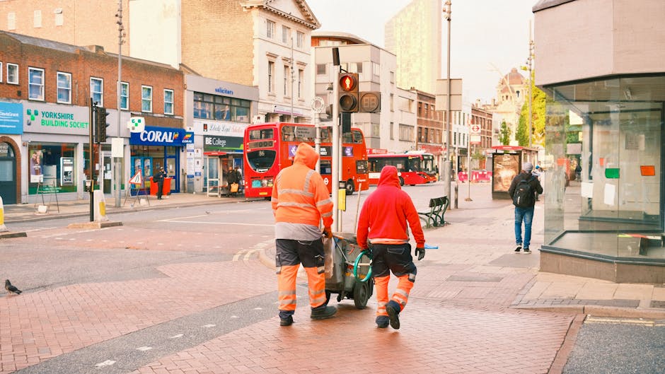 Two workers dressed in high-visibility orange and red safety clothing are seen pushing a wheeled bin or trolley along a busy urban street, possibly engaged in rubbish removal or waste collection. One