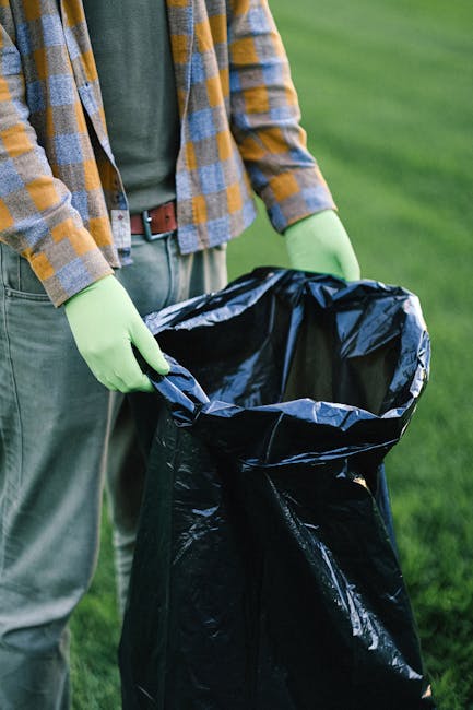 A person wearing a checkered shirt in shades of yellow, blue, and gray, with a green top underneath, is holding open a large black rubbish bag lined with shiny black plastic. The individual is standin